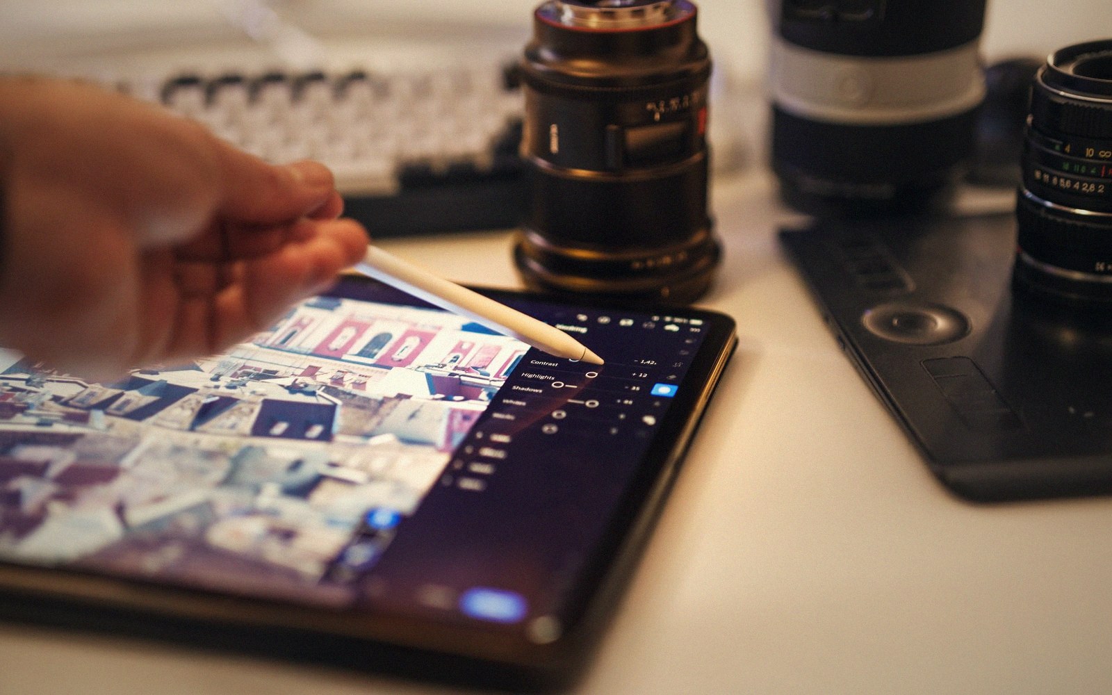 A hand uses a stylus to edit photo contrast settings on a tablet beside camera lenses